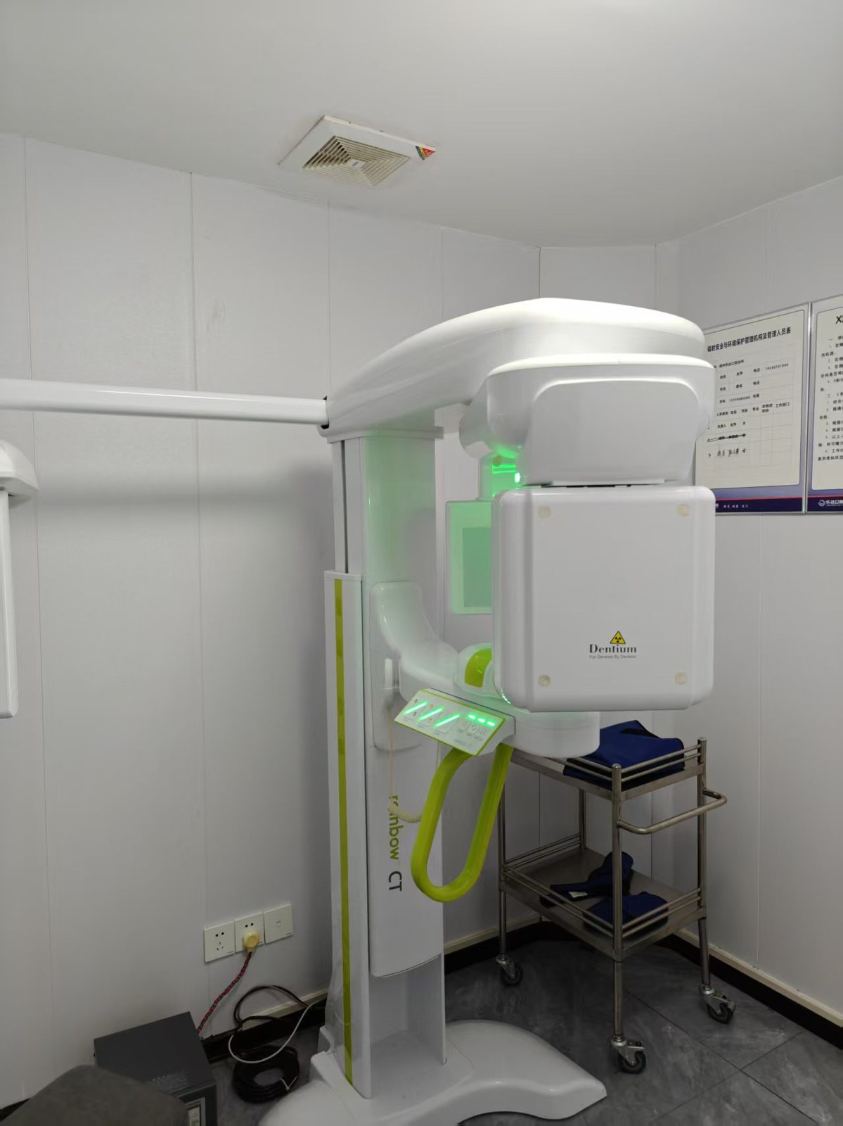 Dentium Rainbow CT photographed in an operational dental radiology room — showing the glowing green status LED indicating standby mode, the control panel with push-button console, lead apron trolley beside the unit, and the radiation safety management notice on the wall