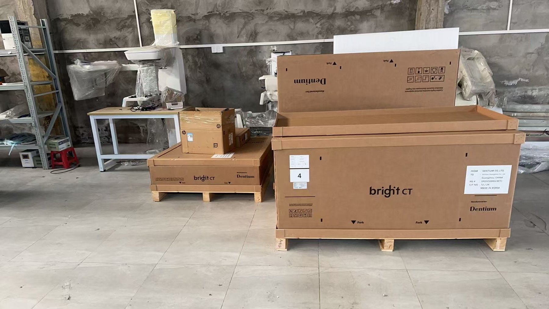 Dentium factory export crates photographed in a distribution warehouse — three wooden crates total per unit, with the main body crate clearly labelled Dentium Manufactured-for and a shipping label showing origin Dentium Co. Ltd. destined for Dentium Guangzhou, China, photographed against a concrete wall with a stocking rack visible in the background